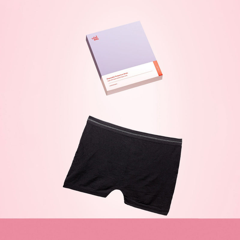 Black disposable postpartum briefs with it's packaging on a pink background