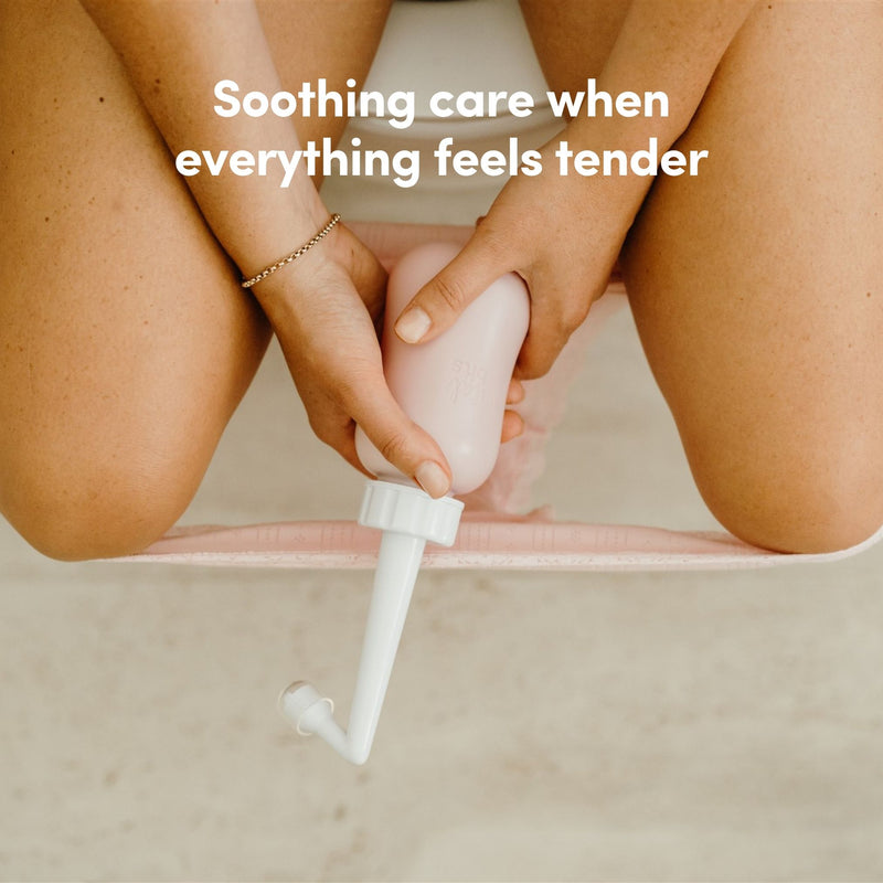 A woman sitting on a toilet holding a peri bottle between her legs with text 'Soothing care when everything feels tender' on a neutral background.