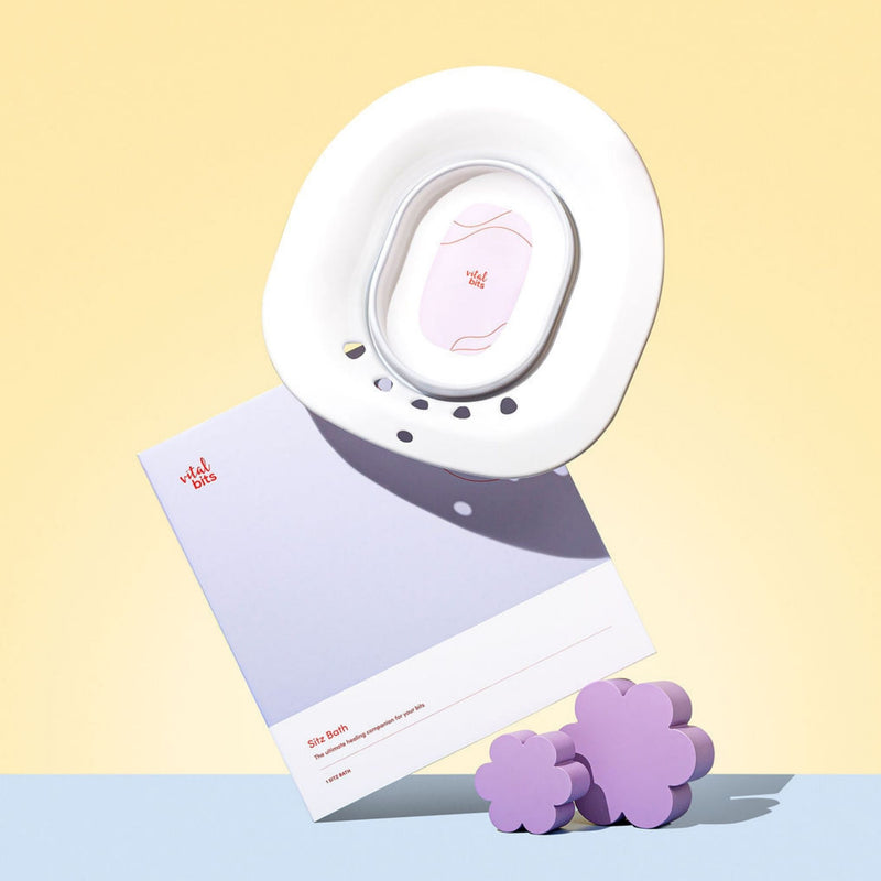 A styled image of a Vital Bits Sitz Bath with its box and design props on a light yellow and blue gradient background. 
